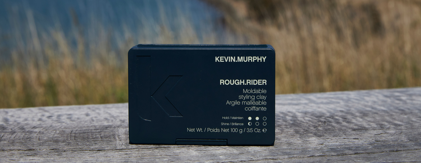 Outdoor image of KEVIN.MURPHY ROUGH.RIDER styling clay packaging, displaying the product's name and attributes like hold and shine level on the label, set against a blurred natural backdrop.